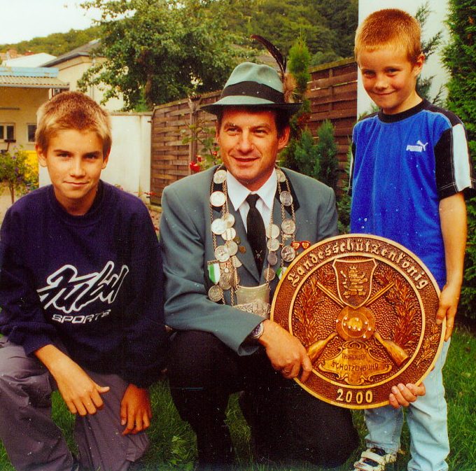 Zwei Jungen stehen neben einem Mann in Anzug, der eine große Ehrenscheibe hält.